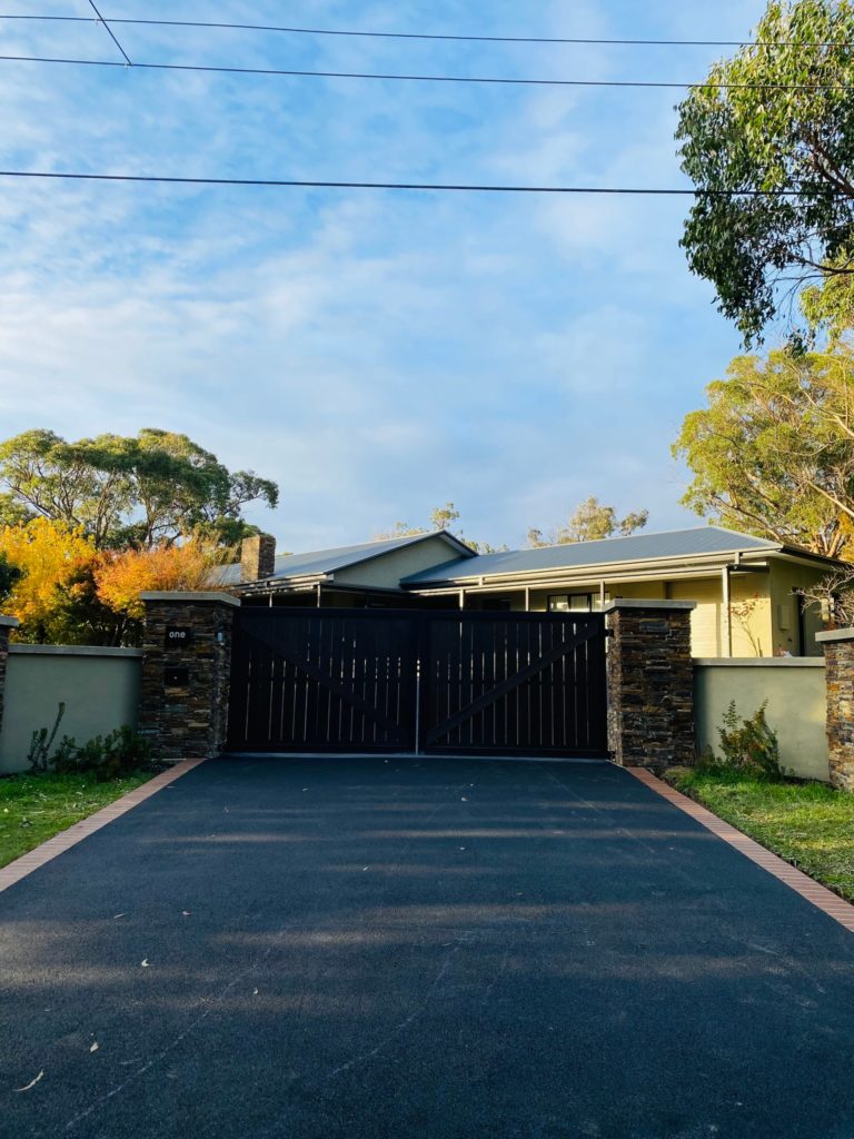 asphalt driveway with stacked stone gateway pillars. The gate is closed but there is a house behind and a blue sky.