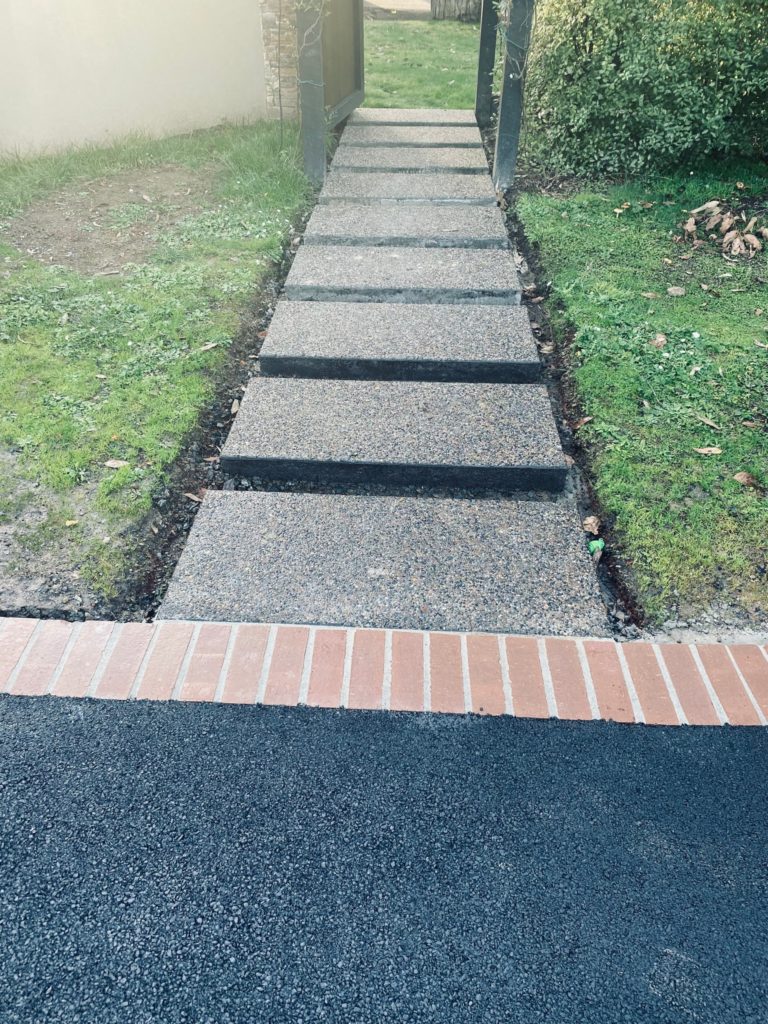 Exposed aggregate stepping stones with bricked edged asphalt in the foreground. The stepping stones lead through an archway.